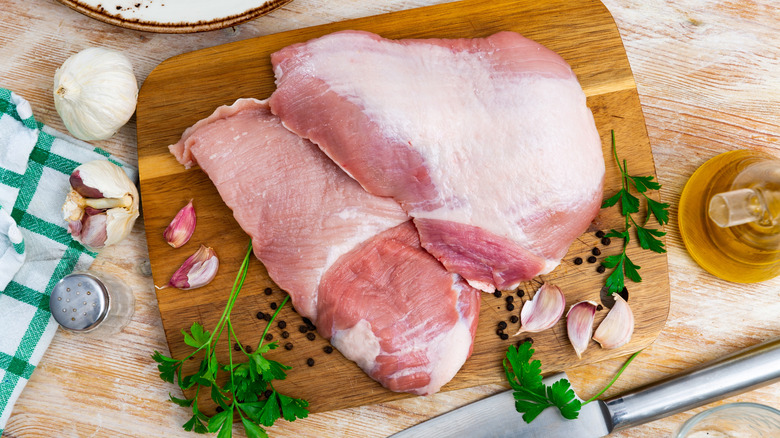 Pieces of raw pork rest on a wooden cutting board with herbs and garlic cloves