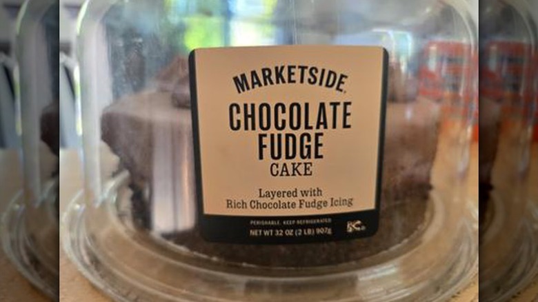 A Marketside Chocolate Fudge Cake sits on a white cake stand next to a slice of the cake
