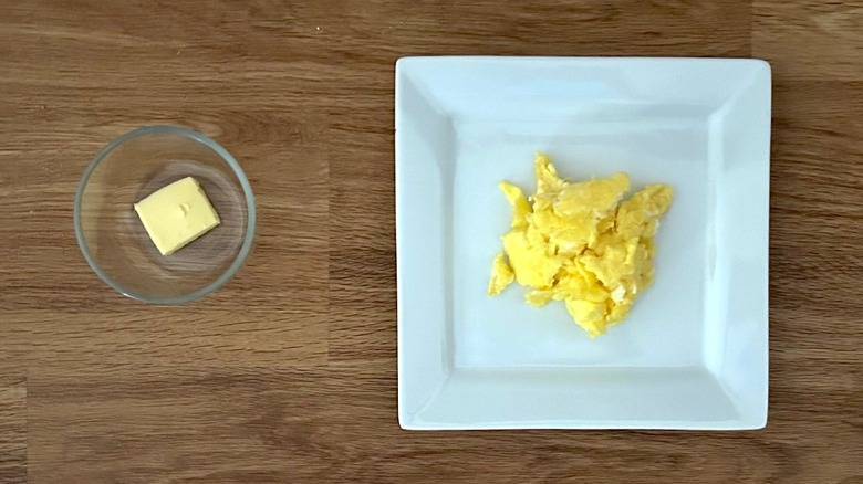 Glass ramekin of butter and square white plate of scrambled eggs