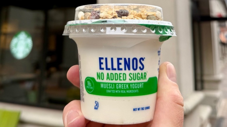 Someone holds up a container of Ellenos Muesli Yogurt in front of a Starbucks