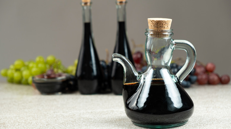 Balsamic glaze in a glass cruet stopped with a cork, and grapes and vinegar in the background.