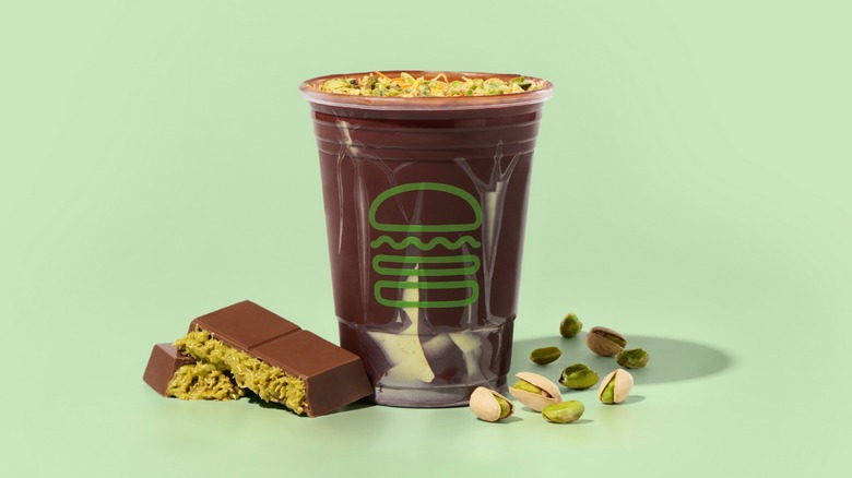 Shake Shack's new Dubai Chocolate Pistachio Shake in a cup next to a bar of Dubai chocolate and some pistachios in and out of their shells