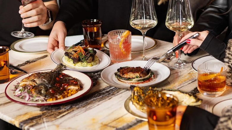 a marble dinner table full of various plates of food and beverages with hands and forks