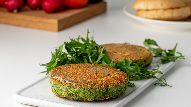 A broccoli-based veggie burger looks pleasantly crispy and rests on a cutting board with some fresh arugula