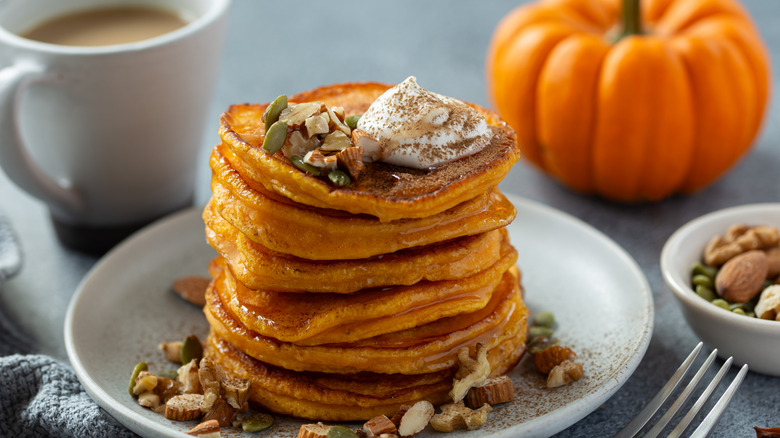 A stack of pancakes stopped with seeds and nuts, with a pumpkin in the background.