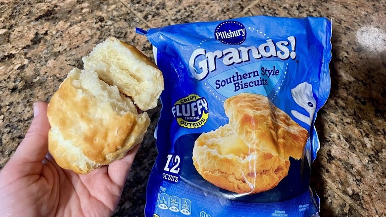 Someone holds a cooked Pillsbury Grands! Southern Style Biscuit next to its packaging, displaying its flaky layers.