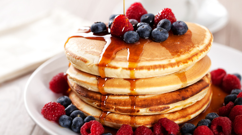 Pancakes on a plate are topped with fresh blueberries and raspberries, with a drizzle of amber syrup