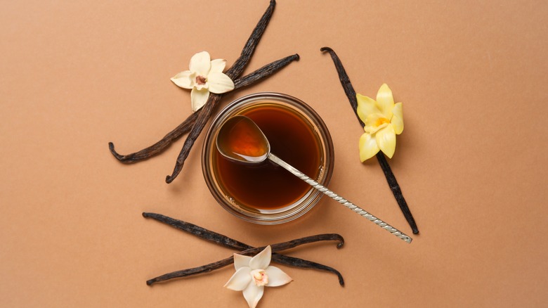 A bowl of vanilla extract surrounded by vanilla pods and flowers