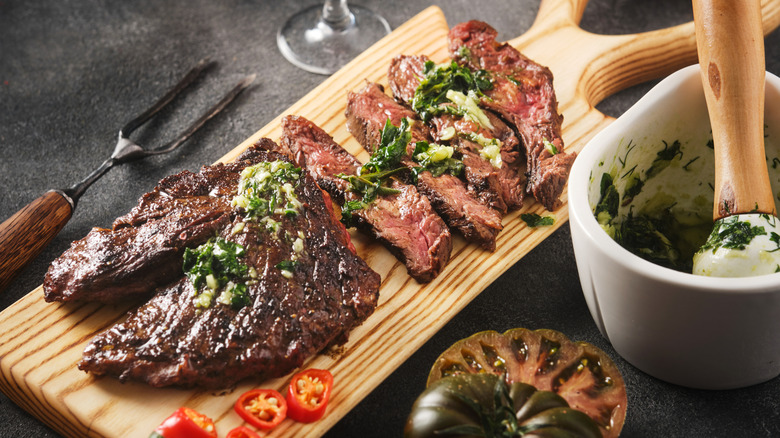 Barbecued hanger steak cut rests on a wooden slab with fresh tomato, chilies, and chimichurri.