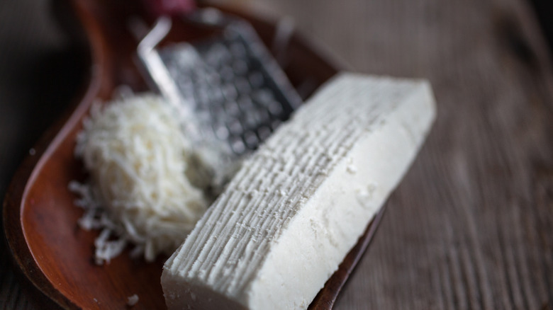 Ricotta salata with markings from a cheese grater. A pile of shredded cheese and the cheese grater are out of focus in the background.