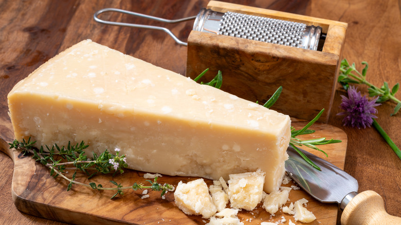A wedge and crumbles of Grana Padano on a wooden cutting board next to fresh herbs and cheese grater
