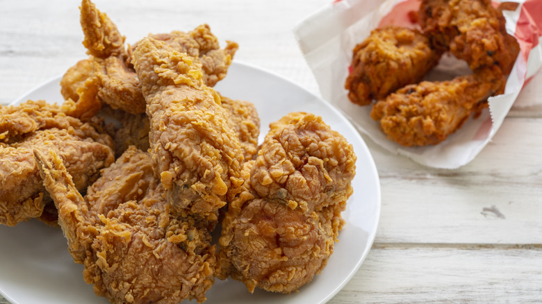 Fried chicken sits on a white plate, with fried drumsticks sitting nearby on a paper wrapper