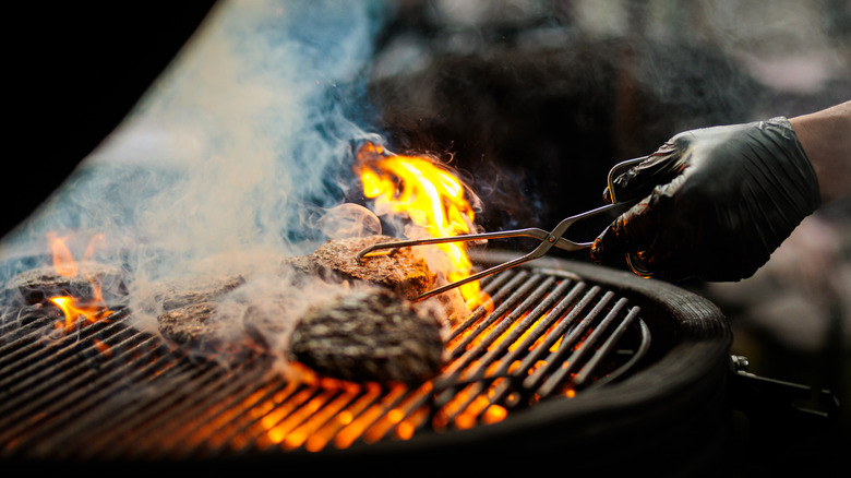 Close-up shot of a gloved hand using tongs to turn hamburgers on a flaming grill.