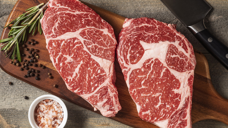 Uncooked wagyu steaks on a wooden cutting board with peppercorns, pink rock salt, and rosemary