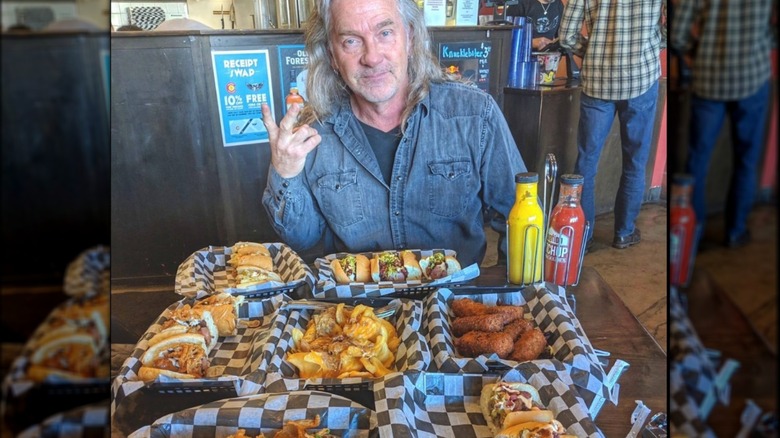 Biker Jim himself sits at a tale in his restaurant, with a wide array of food laid before him, including tater kegs, hot dogs, and more.