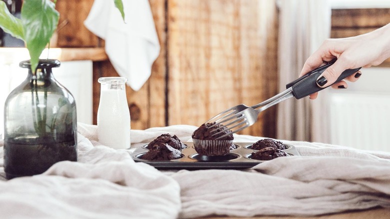 A hand holding Anytongs removes a chocolate muffin from a muffin tin