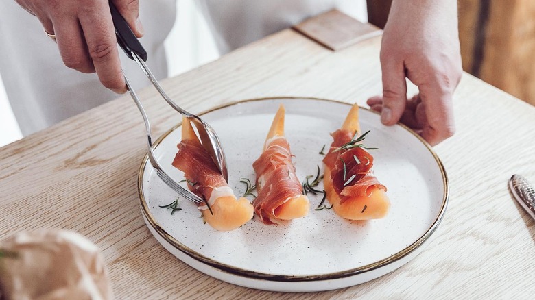 Hands holding Anytongs place a piece of prosciutto-wrapped melon on a plate