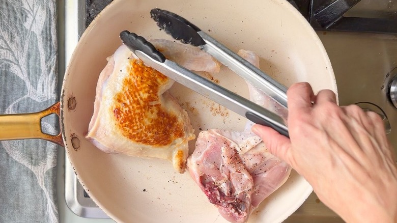 Chicken leg quarters browning in white skillet with tongs