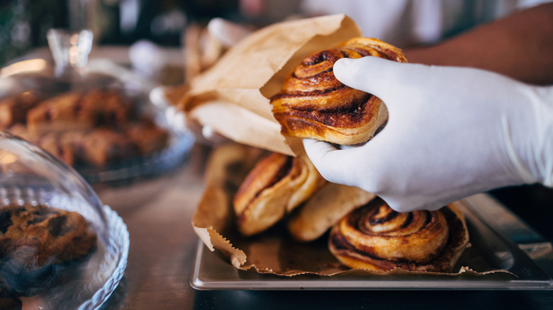 A cinnamon roll is served by a gloved hand in a bakery.