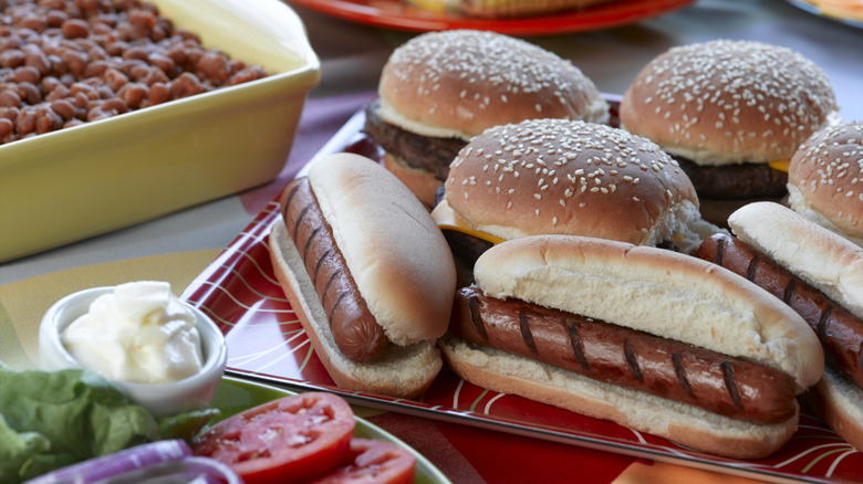 A platter of hot dogs and burgers sit next to condiments and fresh toppings