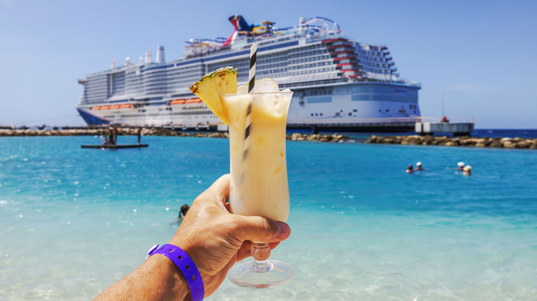 A hand holds up a tropical beverage in front of blue seas and a cruise ship in the background