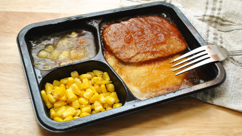 A frozen TV dinner sits on a wooden table next to a silver fork