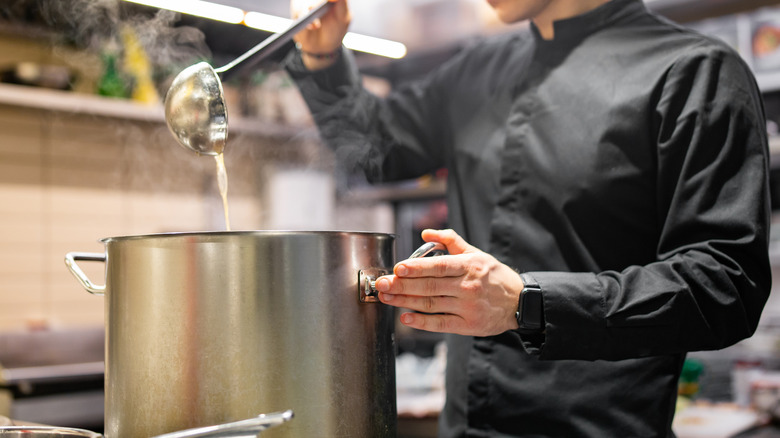 A chef in all-black attire scoops a ladle of soup from a large silver pot