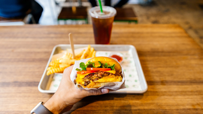 A hand holding a burger with veggies with crinkle-cut fries on a tray in the background