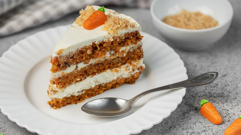 A three-layer piece of carrot cake sits on a white plate with a spoon