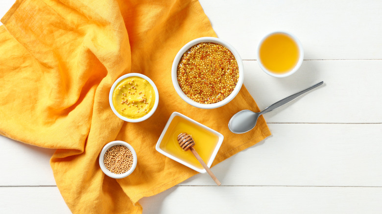 Bowls of mustard and honey sit on an orange towel on a white, wooden surface