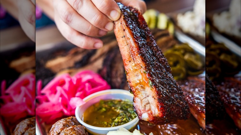 A rib being held over a bbq plate with a dipping sauce and pickled red onions