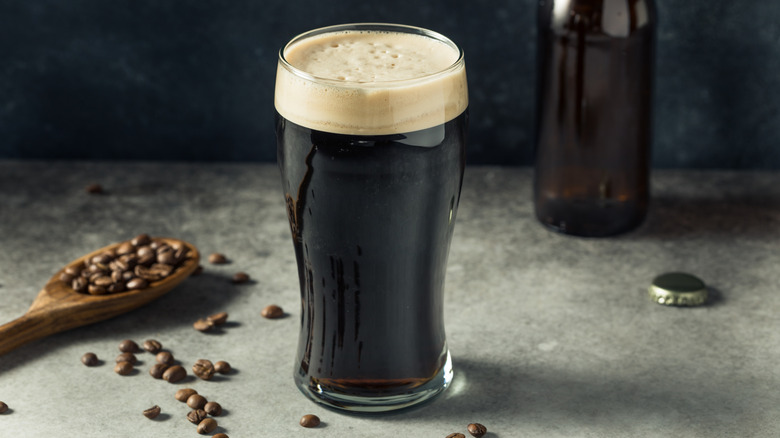 Tall glass of dark beer on gray service with a wooden spoon with coffee beans in the backrgound