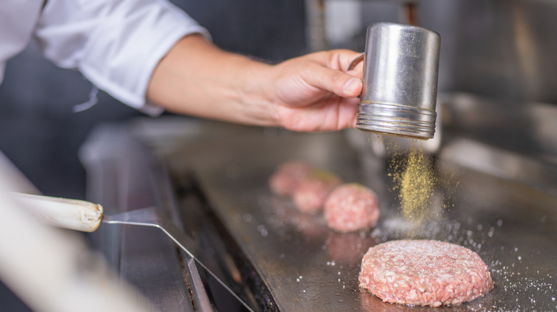 A cook sprinkles seasoning on burger patties on the griddle