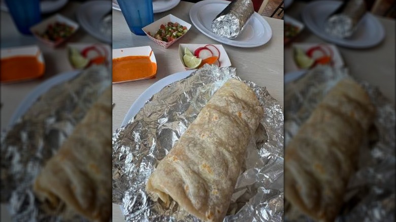A burrito on tin foil on a table across from another burrito with salsa and garnishes.