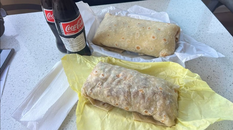 Two burritos on a table next to two bottles of Coke.