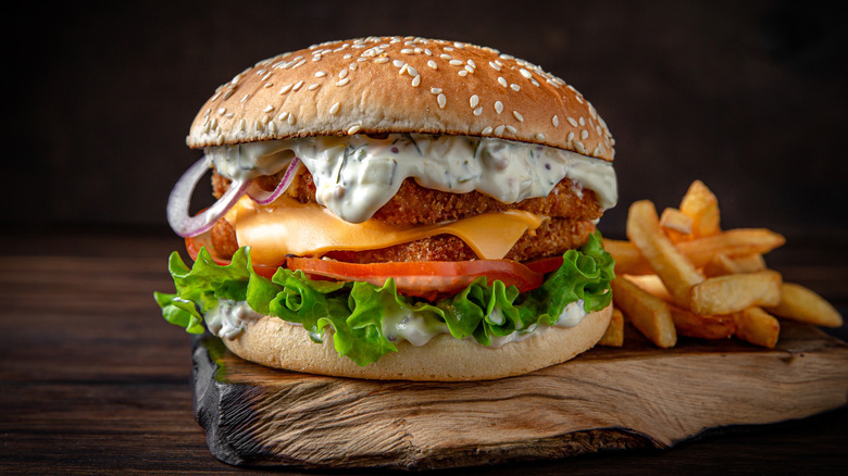 Crispy fish sandwich dripping with tartar sauce served on a sesame seed bun atop a plank with fries