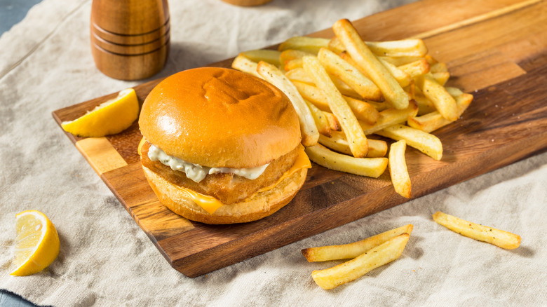 A fried fish sandwich with tartar sauce and fries rest on a wooden plank.