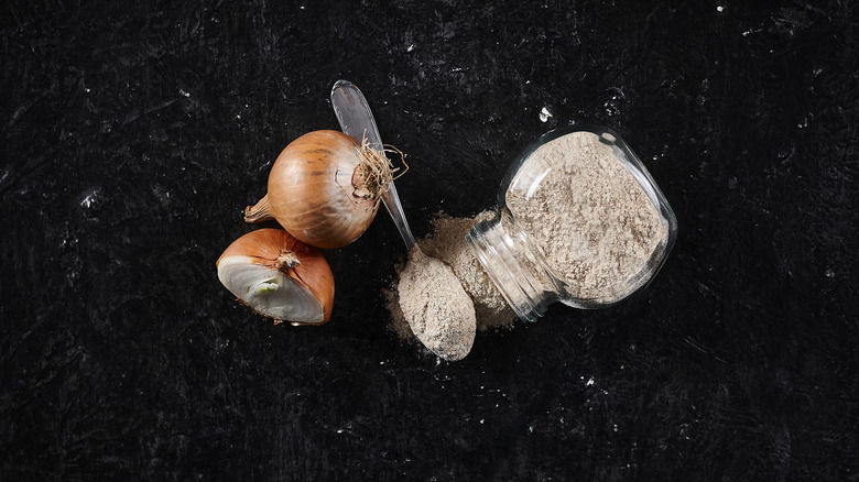 Onion, spoon, and glass jar of onion powder on its side with some spilling out