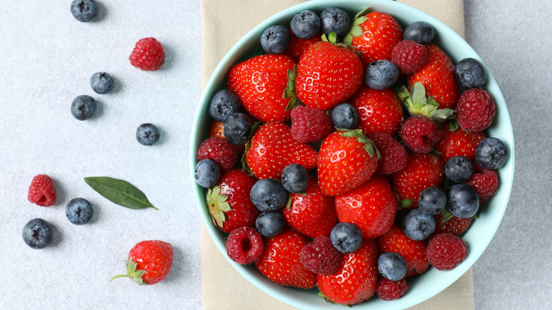 A bowl of strawberries, blueberries, and raspberries with berries scattered adjacent.