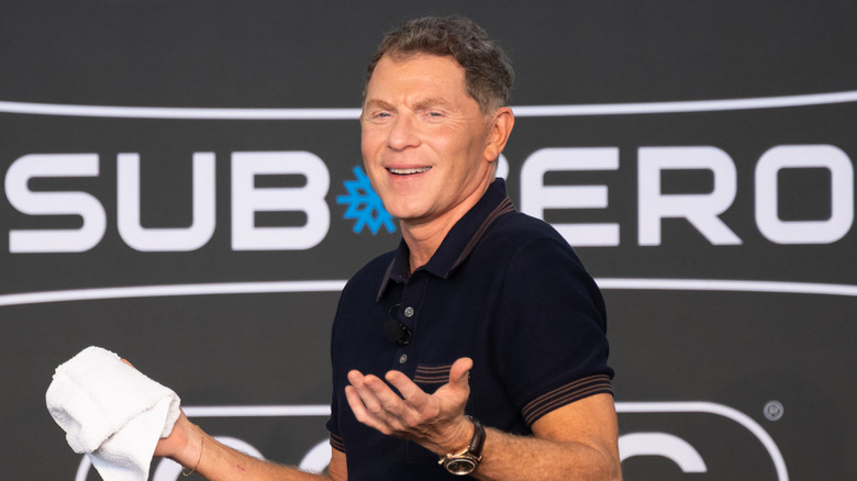 Bobby Flay shrugs while smiling at a cooking demonstration.