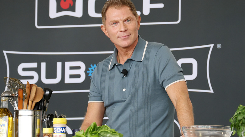 A smiling Bobby Flay stands at a cooking station that is covered with ingredients and cooking tools