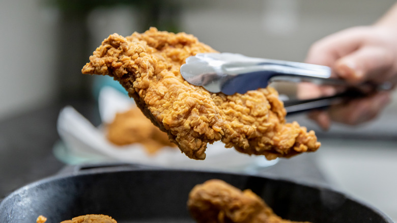 Tongs hold a piece of fried chicken.