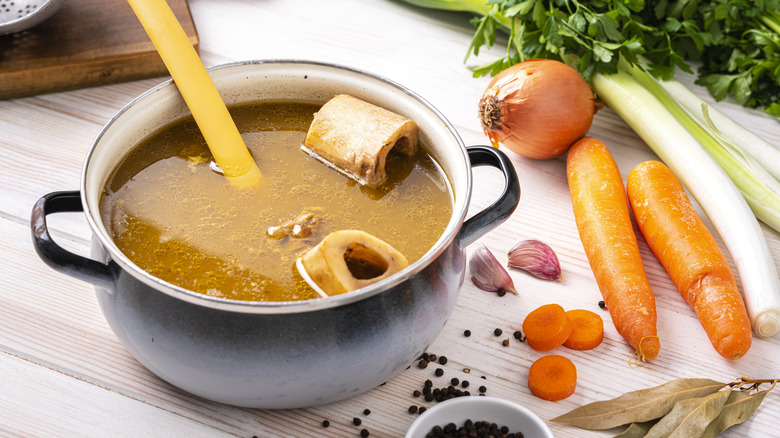 pot of bone broth with ladle and carrots, celery, garlic, and peppercorns