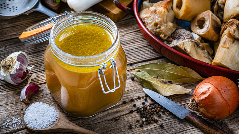 mason jar of bone broth with countertop full of garlic, onions, bay leaves, bones, salt, and peppercorns