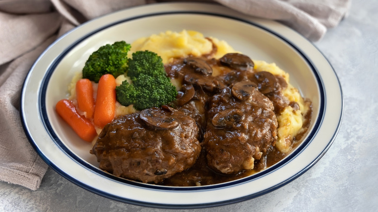 Plate with Salisbury steak topped with mushrooms and brown gravy with mashed potatoes, carrots, and broccoli behind it on the plate
