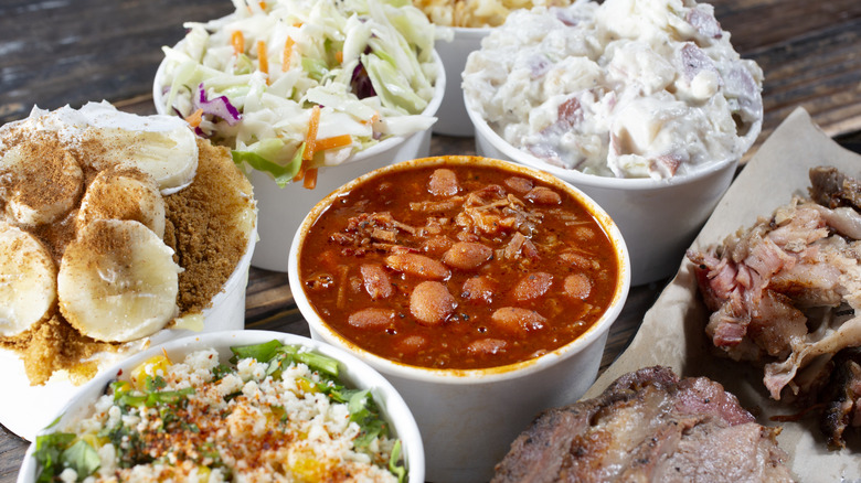 A side of baked beans along with other barbecue sides such as coleslaw, banana pudding, salad, smoked meat, and potato salad.