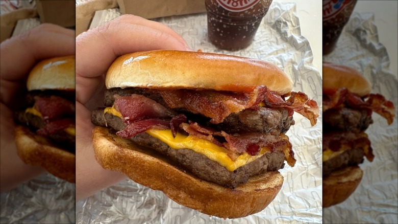 A hand holds up a Wendy's Baconator, with the burger wrapper and a Wendy's soda in the background