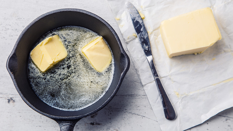 Two pats of butter melting in a dark pan next to knife and butter stick