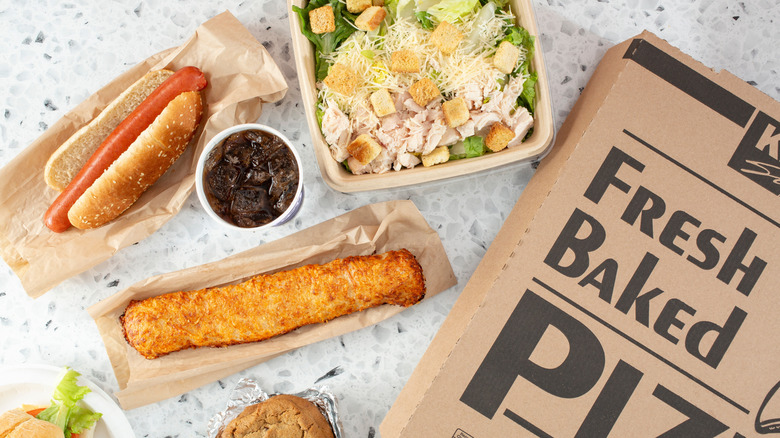 A selection of Costco food court food items laid out on a table including a caesar salad, hot dog, chicken bake, cookie, sandwich, soda, and a box of pizza