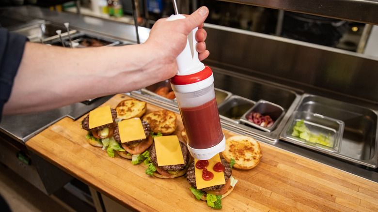 A hand uses a fast food sauce gun full of ketchup to dress open cheeseburgers